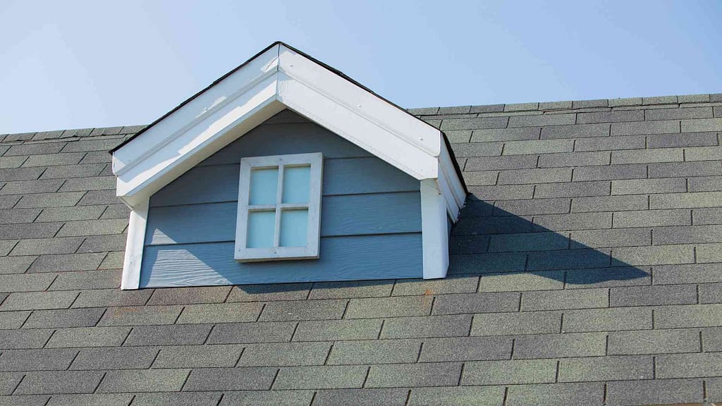 Image of a roof with gray shingles, blue house color, and a small square window, installed by American Home Contractors in Green Village, NJ.