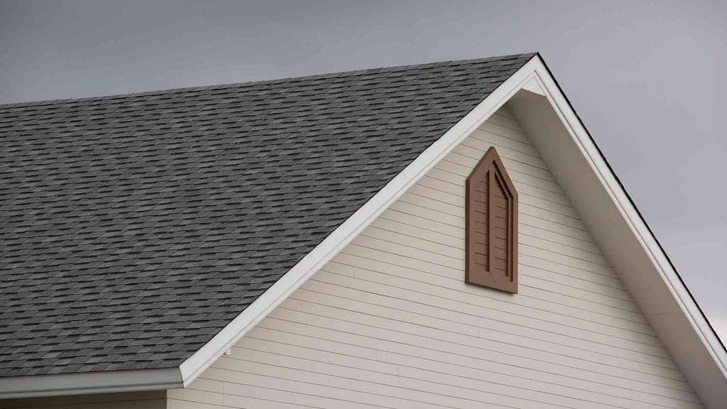 Image of a light tan house with dark gray asphalt shingles and white trim, installed by Roofing Contractor in Madison, NJ, against a gray sky background.
