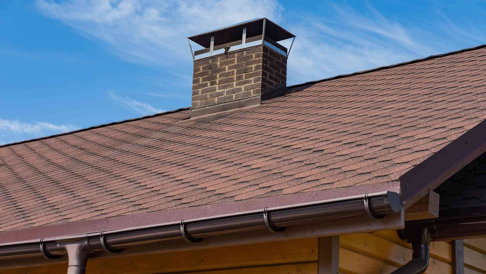 Image of a house roof with brown shingles and a brick chimney, installed by professional Roofing Contractors in Madison, NJ.